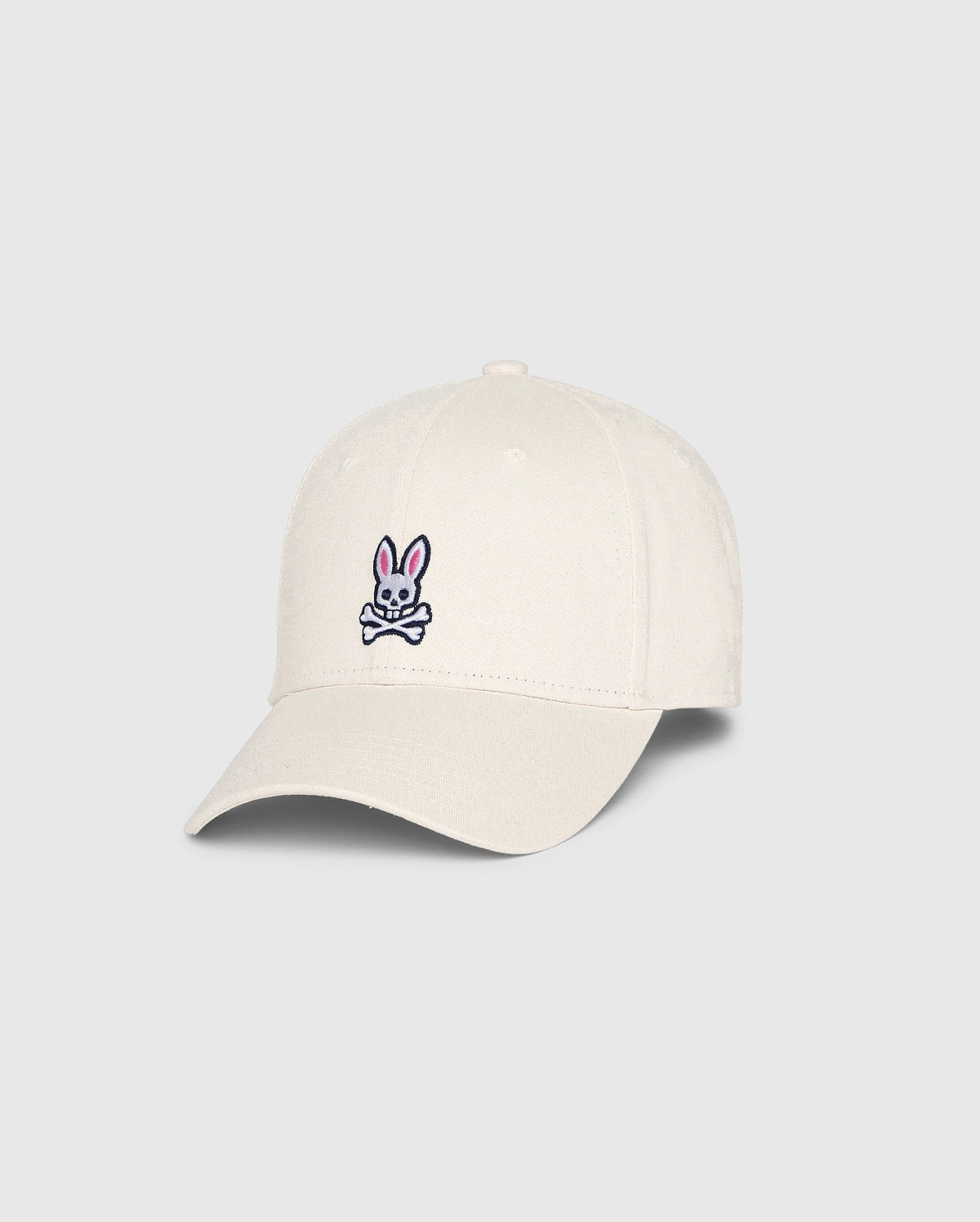 The Psycho Bunny MENS CLASSIC BASEBALL CAP (B6A816B200), made from 100% cotton in beige, showcases a small pink bunny with crossed bones on the front. Angled to highlight both front and side, this standout accessory adds character to any collection.