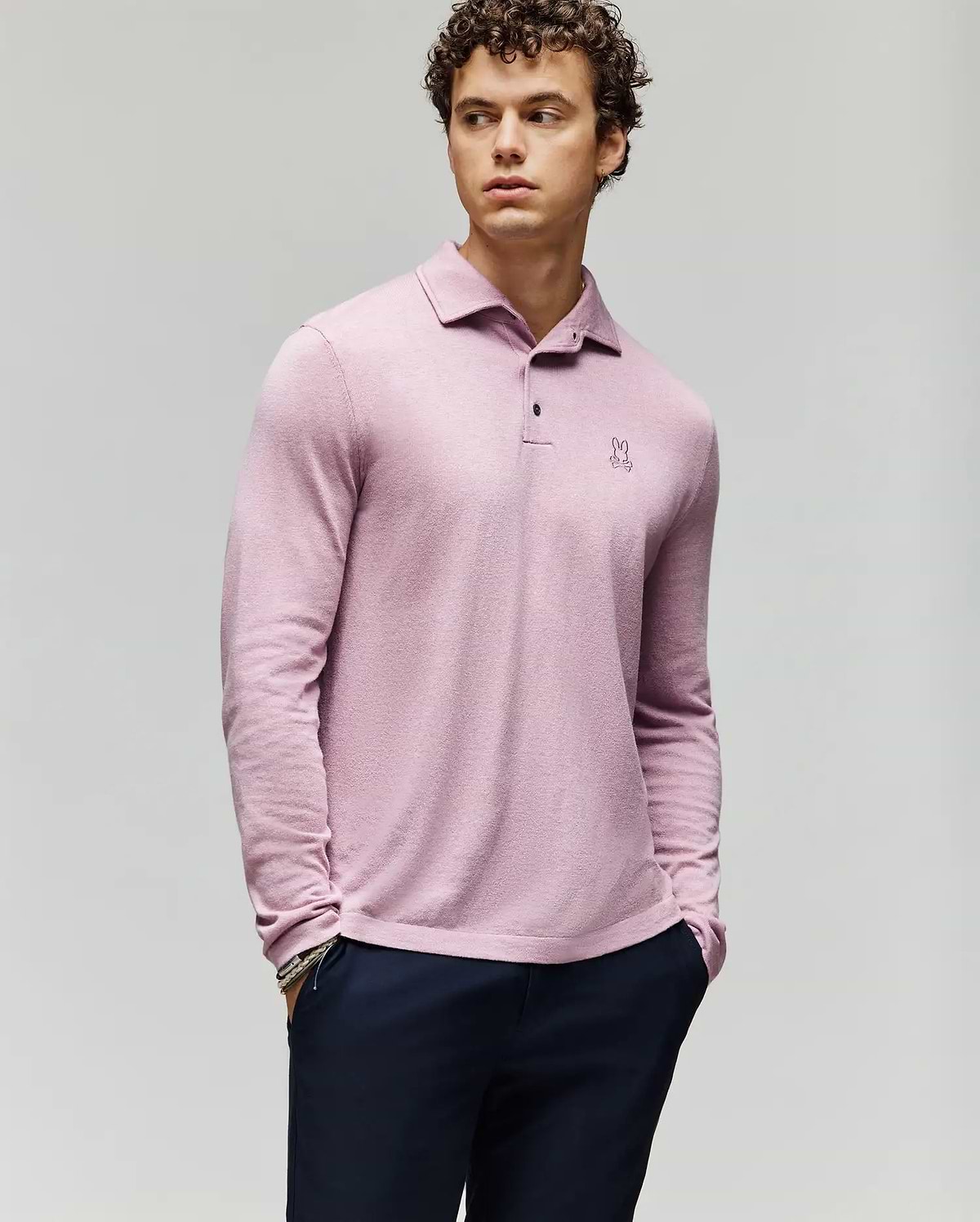 A young man with curly hair wears the Psycho Bunny MENS BRADSHAW POLO SWEATER (B6E835D200) in light pink, featuring a small embroidered logo and mother of pearl buttons, paired with dark pants against a light gray background. (1)