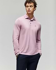 A young man with curly hair wears the Psycho Bunny MENS BRADSHAW POLO SWEATER (B6E835D200) in light pink, featuring a small embroidered logo and mother of pearl buttons, paired with dark pants against a light gray background. (1)