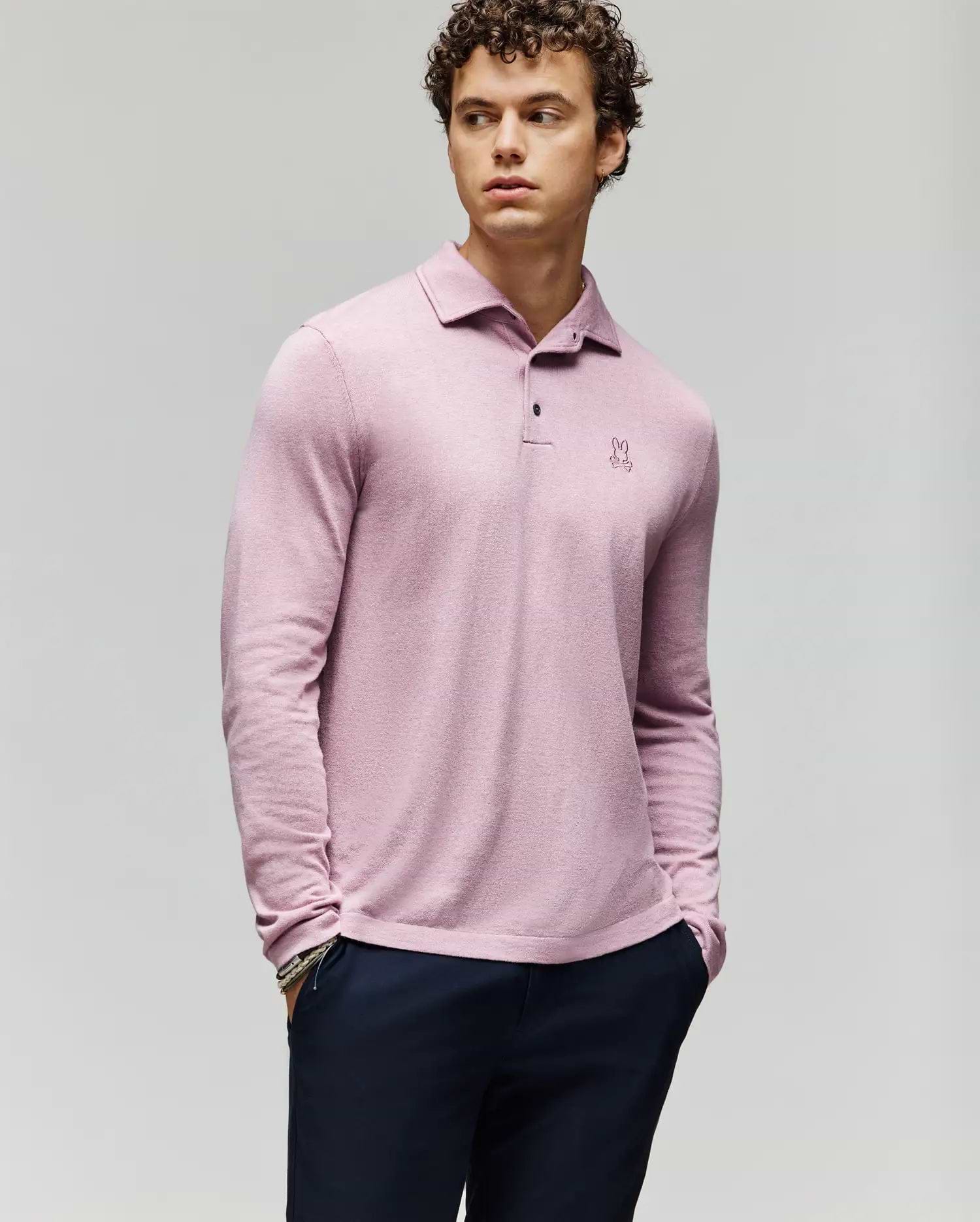 A young man with curly hair wears the Psycho Bunny MENS BRADSHAW POLO SWEATER (B6E835D200) in light pink, featuring a small embroidered logo and mother of pearl buttons, paired with dark pants against a light gray background. (1)