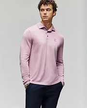 A young man with curly hair wears the Psycho Bunny MENS BRADSHAW POLO SWEATER (B6E835D200) in light pink, featuring a small embroidered logo and mother of pearl buttons, paired with dark pants against a light gray background. (1)