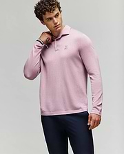 A man with curly hair wears the Psycho Bunny MENS BRADSHAW POLO SWEATER (B6E835D200) in light pink with mother of pearl buttons and dark pants, posing with one hand on his neck against a plain light background. (3)