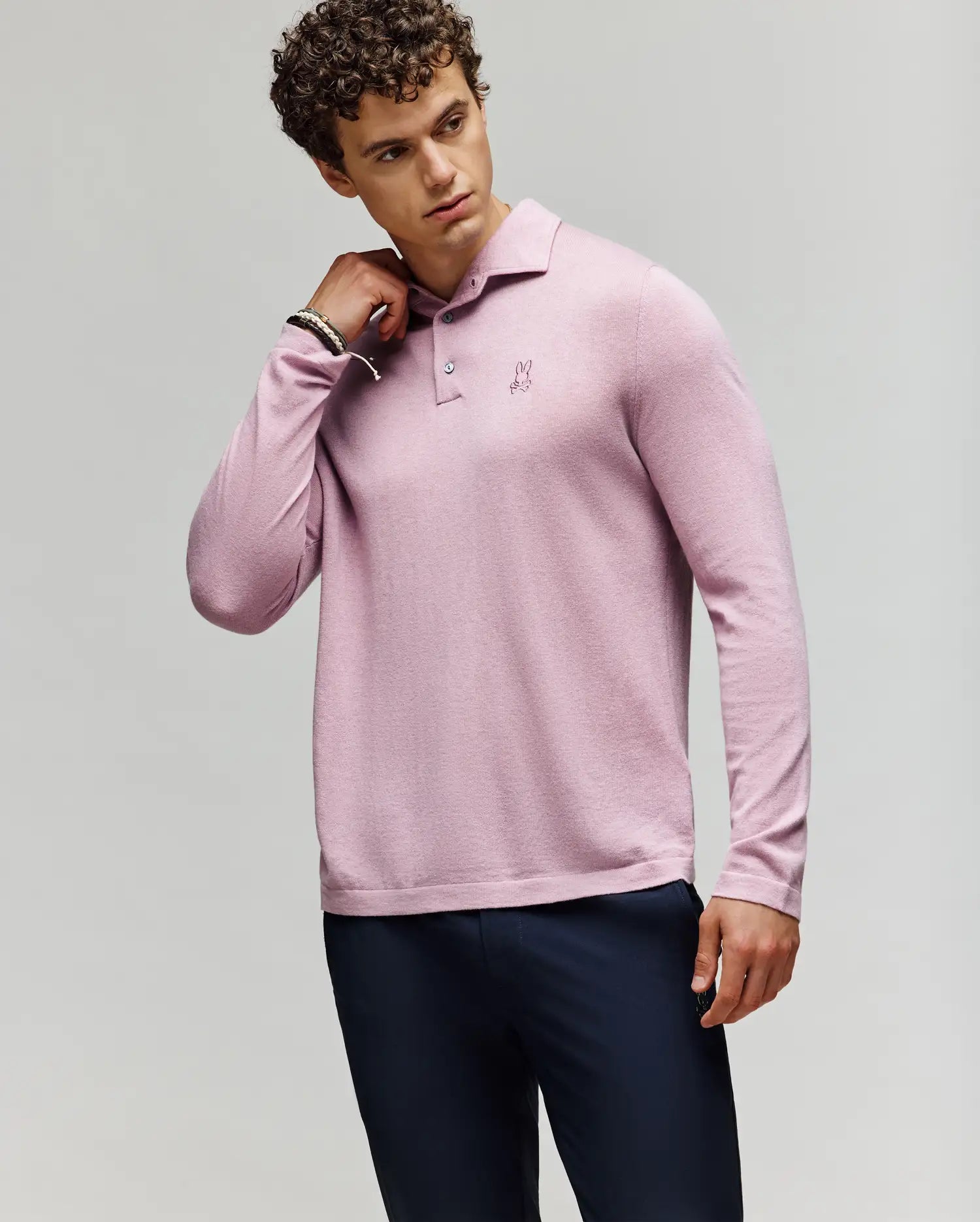 A man with curly hair wears the Psycho Bunny MENS BRADSHAW POLO SWEATER (B6E835D200) in light pink with mother of pearl buttons and dark pants, posing with one hand on his neck against a plain light background. (3)