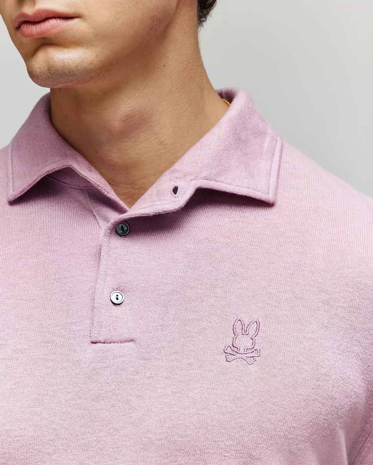 A man wears the Psycho Bunny MENS BRADSHAW POLO SWEATER (B6E835D200) in light purple, featuring a small embroidered bunny and crossbones logo on the chest and mother of pearl buttons against a neutral background. (2)