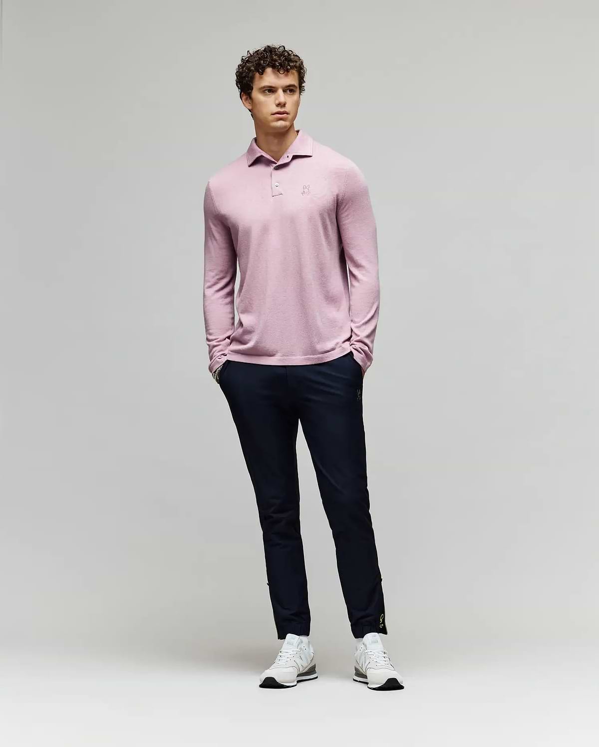 A man with curly hair stands against a plain background wearing the Psycho Bunny MENS BRADSHAW POLO SWEATER - B6E835D200 in light pink, paired with dark navy pants and white sneakers. His hands are in his pockets as he looks slightly to the side. (4)