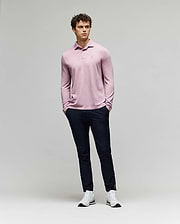A man with curly hair stands against a plain background wearing the Psycho Bunny MENS BRADSHAW POLO SWEATER - B6E835D200 in light pink, paired with dark navy pants and white sneakers. His hands are in his pockets as he looks slightly to the side. (4)