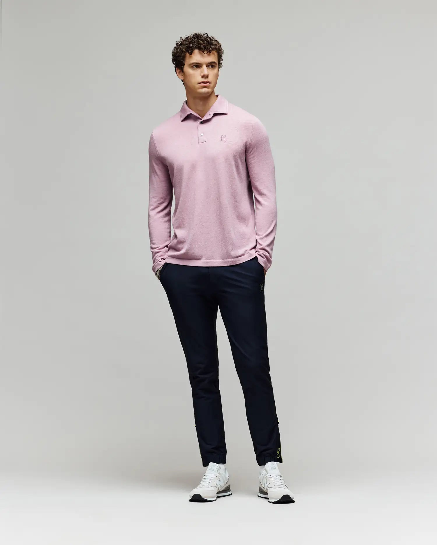 A man with curly hair stands against a plain background wearing the Psycho Bunny MENS BRADSHAW POLO SWEATER - B6E835D200 in light pink, paired with dark navy pants and white sneakers. His hands are in his pockets as he looks slightly to the side. (4)