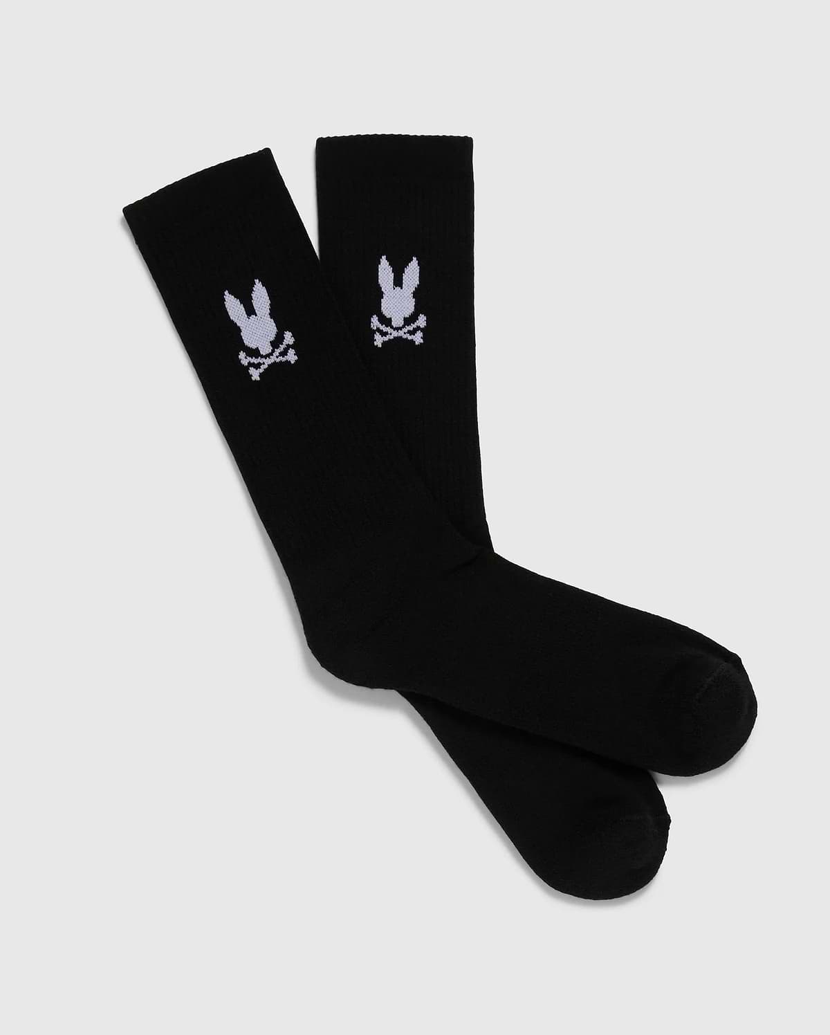 A pack of 3 pairs of black men's knee-high sport socks from the Psycho Bunny brand, featuring a white skull and crossbones symbol with bunny ears on each sock. Made from luxurious Peruvian pima cotton, these MENS 3 PACK SPORT SOCK - B6F997D200 socks are displayed positioned overlapping each other against a plain, light gray background.