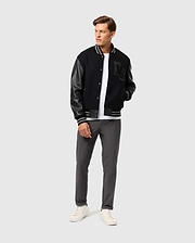 A man wearing the Psycho Bunny MENS BRAYDEN WOOL LEATHER VARSITY BOMBER - B6J876D200, featuring leather sleeves, pairs it with a white t-shirt, gray pants, and white sneakers as he stands against a plain background. With his hands in his jacket pockets, he gazes to the side.