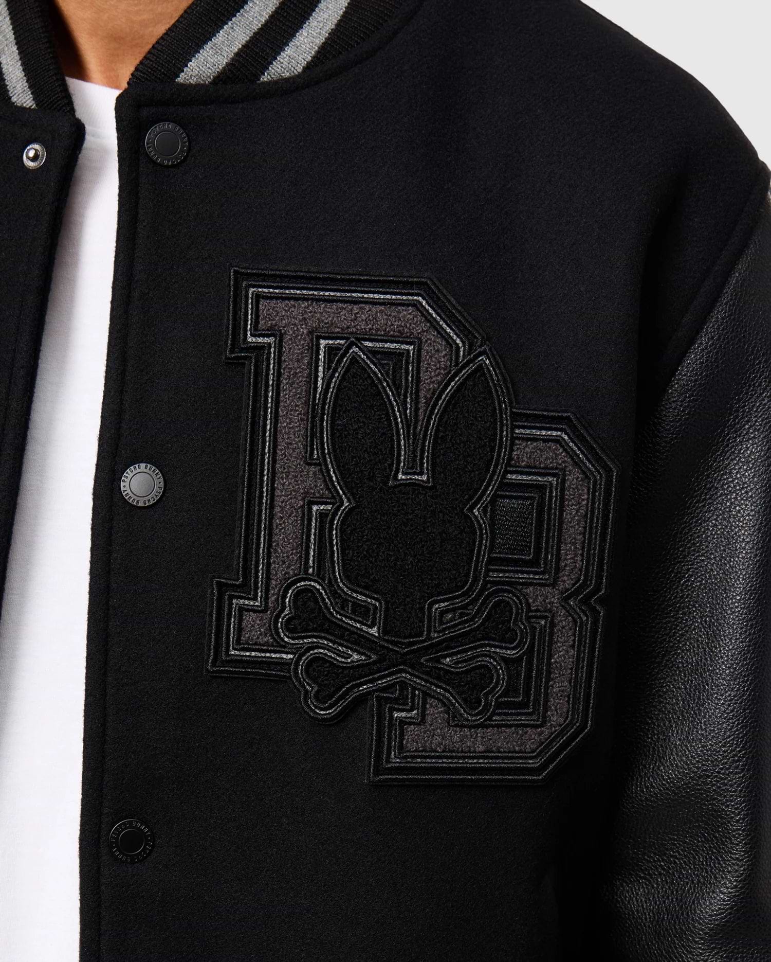 Close-up of the Psycho Bunny Men's Brayden Wool Leather Varsity Bomber (B6J876D200) in black, showcasing an embroidered design featuring large overlapping "DB" letters with a bunny head and crossed bones on the left chest. This wool-blend jacket includes snap buttons, leather sleeves, and a striped collar.