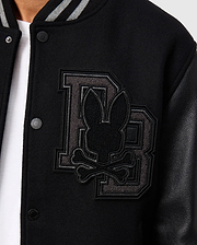 Close-up of the Psycho Bunny Men's Brayden Wool Leather Varsity Bomber (B6J876D200) in black, showcasing an embroidered design featuring large overlapping "DB" letters with a bunny head and crossed bones on the left chest. This wool-blend jacket includes snap buttons, leather sleeves, and a striped collar.