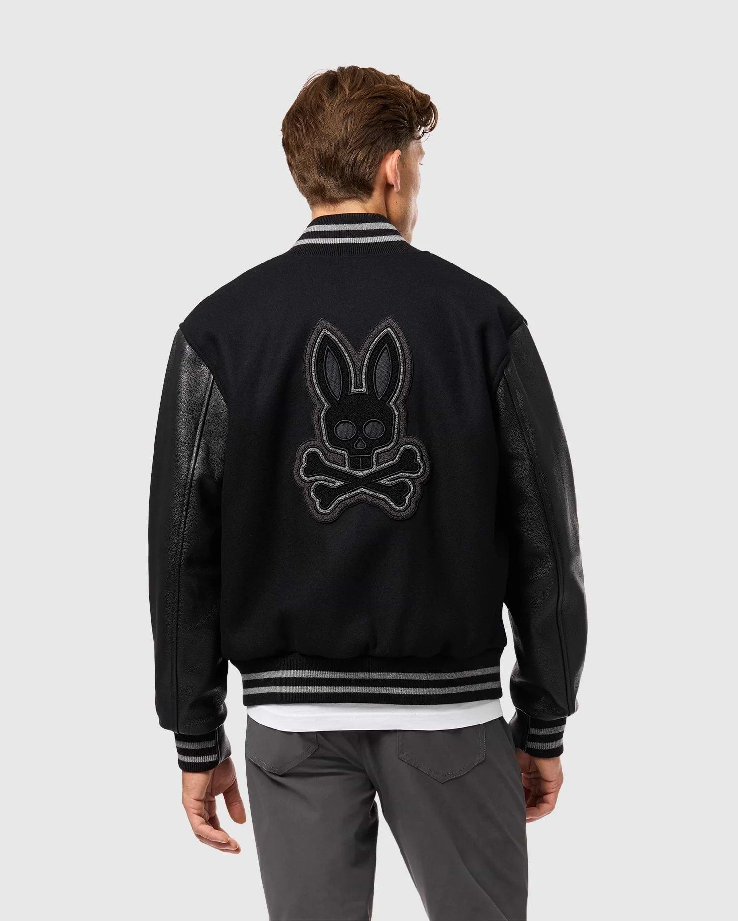 The individual sports the Psycho Bunny MENS BRAYDEN WOOL LEATHER VARSITY BOMBER - B6J876D200, featuring sleek leather sleeves. The back displays a striking skull-and-crossbones motif adorned with rabbit ears. With short brown hair, they're turned away, stylishly pairing the jacket with grey pants and a white shirt beneath.