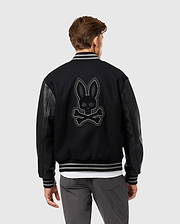 The individual sports the Psycho Bunny MENS BRAYDEN WOOL LEATHER VARSITY BOMBER - B6J876D200, featuring sleek leather sleeves. The back displays a striking skull-and-crossbones motif adorned with rabbit ears. With short brown hair, they're turned away, stylishly pairing the jacket with grey pants and a white shirt beneath.