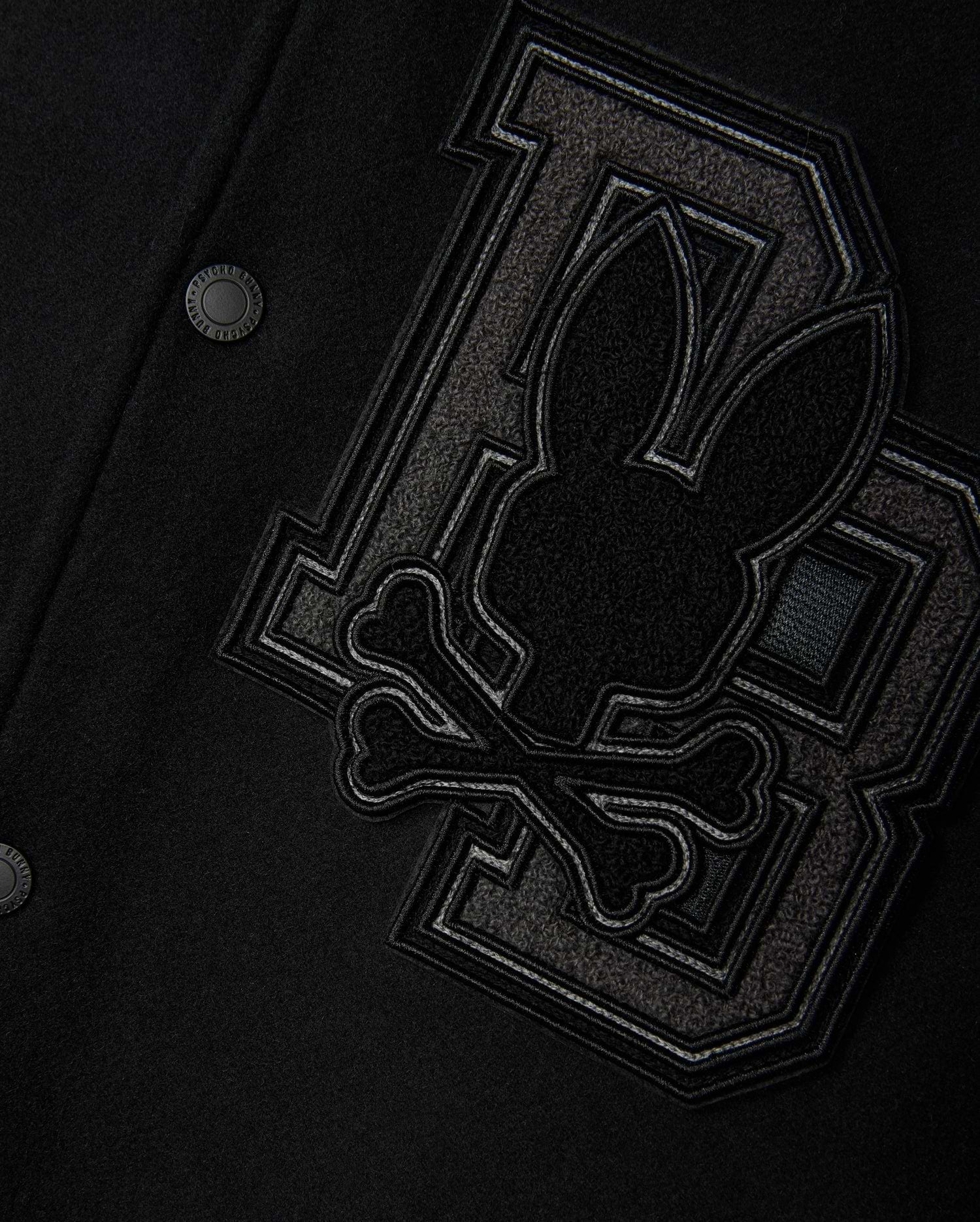 Close-up of the Psycho Bunny Men's Brayden Wool Leather Varsity Bomber - B6J876D200 in black, featuring leather sleeves and a prominent patch. The patch showcases a stylized rabbit head above crossbones, positioned against two large interlocking "BB" letters crafted from textured fabric. This wool-blend jacket is finished with metal snap buttons.