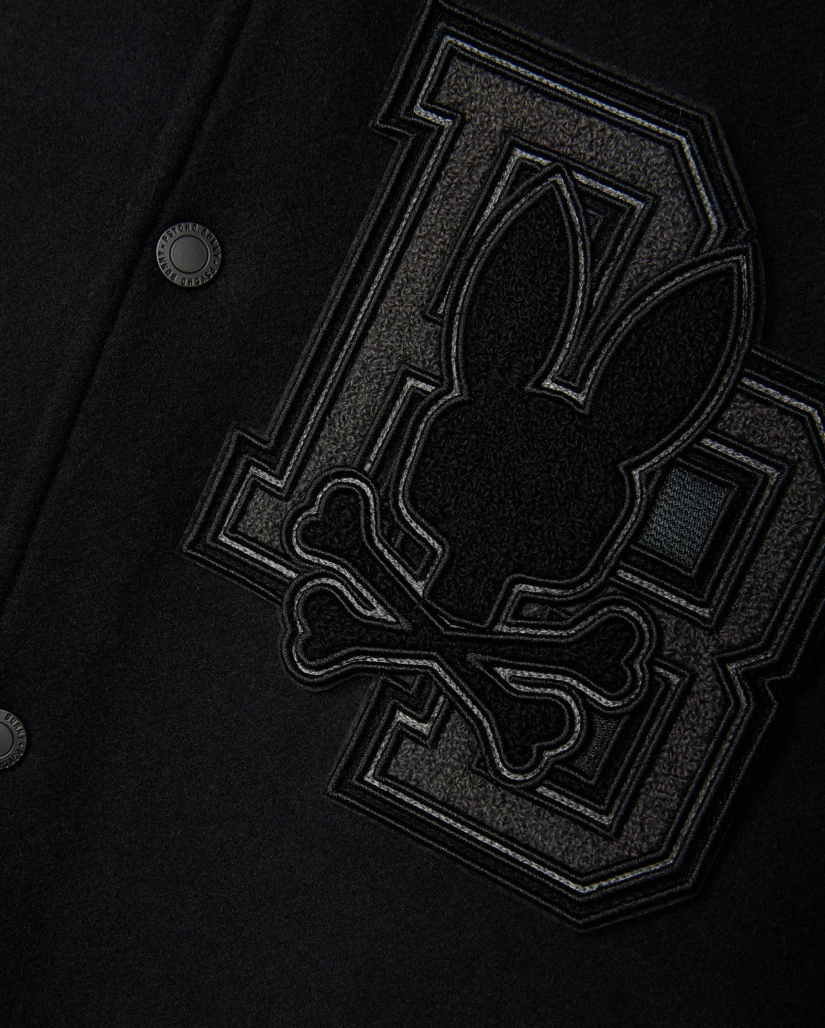 Close-up of the Psycho Bunny Men's Brayden Wool Leather Varsity Bomber - B6J876D200 in black, featuring leather sleeves and a prominent patch. The patch showcases a stylized rabbit head above crossbones, positioned against two large interlocking "BB" letters crafted from textured fabric. This wool-blend jacket is finished with metal snap buttons.