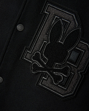 Close-up of the Psycho Bunny Men's Brayden Wool Leather Varsity Bomber - B6J876D200 in black, featuring leather sleeves and a prominent patch. The patch showcases a stylized rabbit head above crossbones, positioned against two large interlocking "BB" letters crafted from textured fabric. This wool-blend jacket is finished with metal snap buttons.