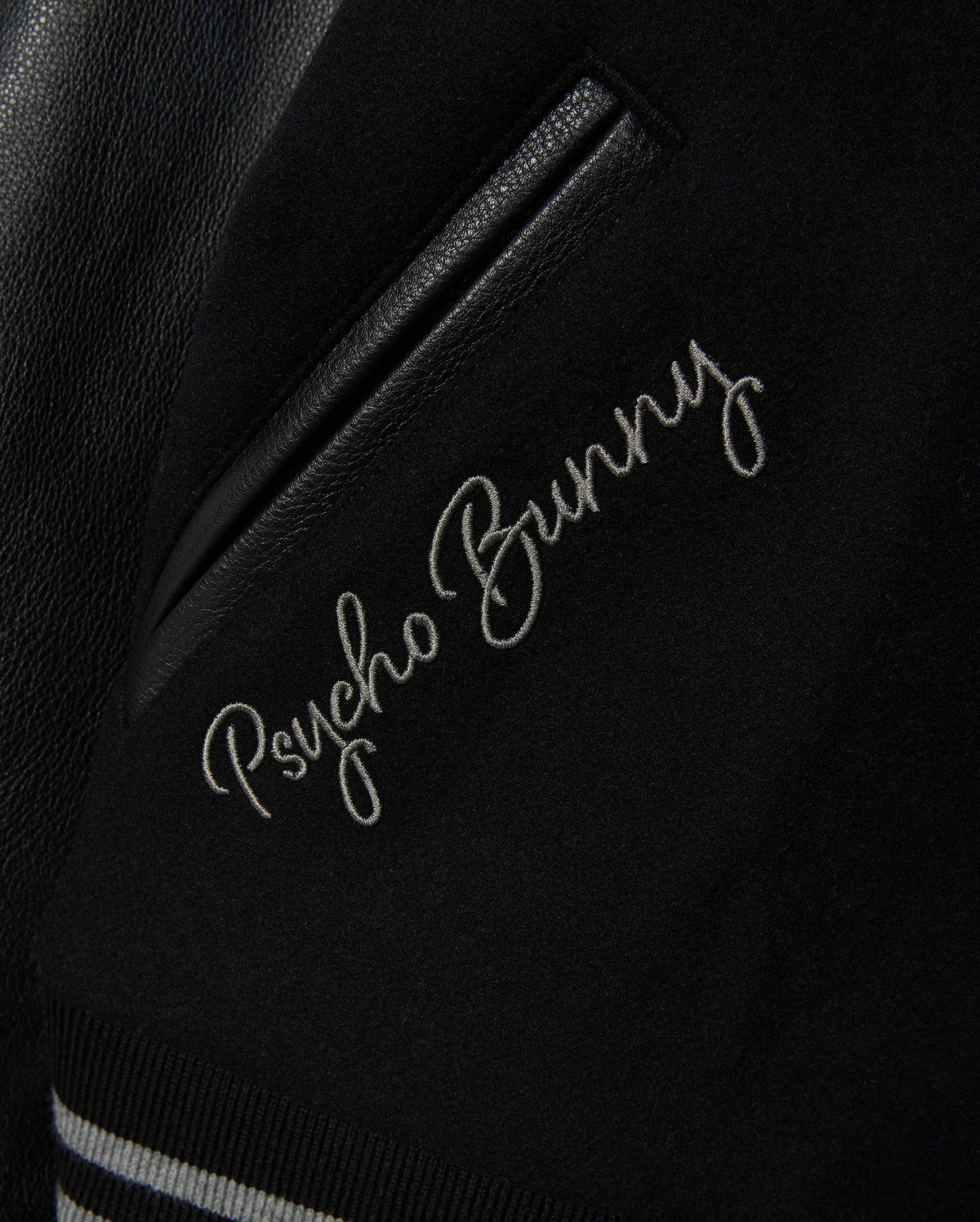 Detailed view of the MENS BRAYDEN WOOL LEATHER VARSITY BOMBER jacket, featuring "Psycho Bunny" elegantly embroidered in white cursive. This black bomber includes leather sleeves, a leather accent, and striped ribbed trim.