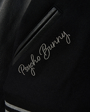 Detailed view of the MENS BRAYDEN WOOL LEATHER VARSITY BOMBER jacket, featuring "Psycho Bunny" elegantly embroidered in white cursive. This black bomber includes leather sleeves, a leather accent, and striped ribbed trim.
