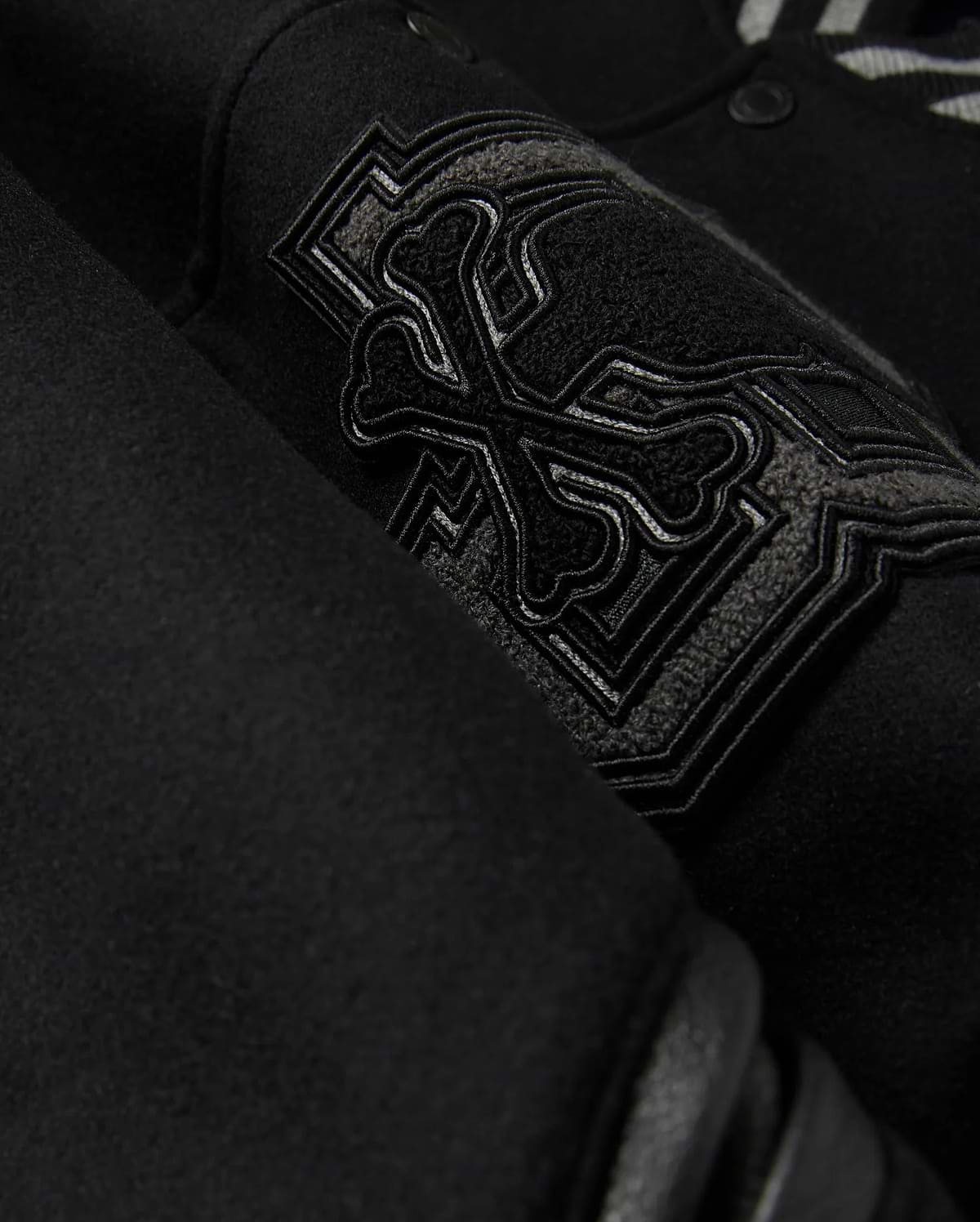 A detailed close-up of the MENS BRAYDEN WOOL LEATHER VARSITY BOMBER - B6J876D200 by Psycho Bunny showcases a black wool-blend fabric with an intricate embroidered design featuring crossbones and decorative elements. The raised and detailed stitching adds texture against the smooth background, reflecting the exceptional craftsmanship typical of a varsity bomber.
