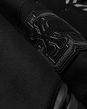 A detailed close-up of the MENS BRAYDEN WOOL LEATHER VARSITY BOMBER - B6J876D200 by Psycho Bunny showcases a black wool-blend fabric with an intricate embroidered design featuring crossbones and decorative elements. The raised and detailed stitching adds texture against the smooth background, reflecting the exceptional craftsmanship typical of a varsity bomber.