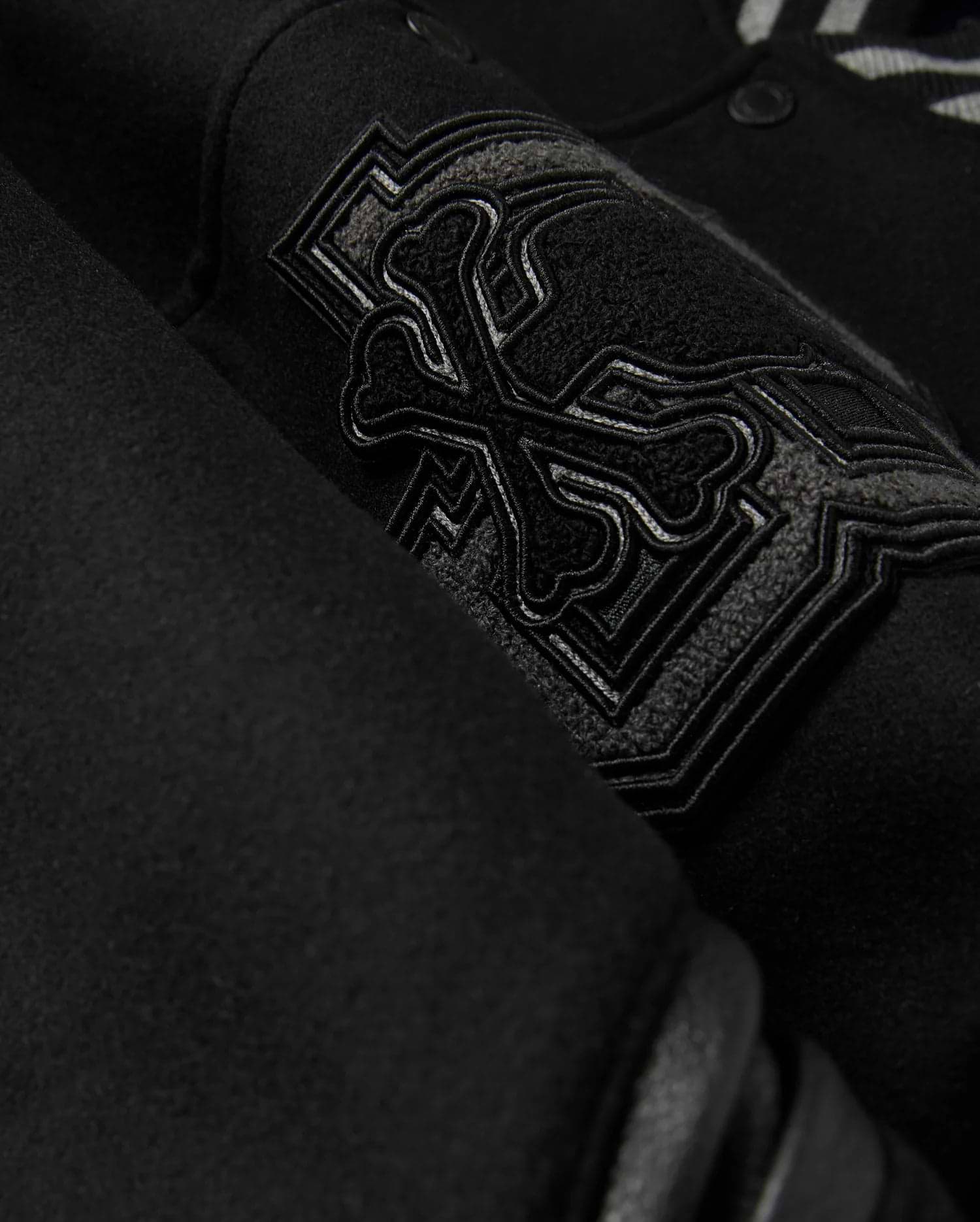 A detailed close-up of the MENS BRAYDEN WOOL LEATHER VARSITY BOMBER - B6J876D200 by Psycho Bunny showcases a black wool-blend fabric with an intricate embroidered design featuring crossbones and decorative elements. The raised and detailed stitching adds texture against the smooth background, reflecting the exceptional craftsmanship typical of a varsity bomber.