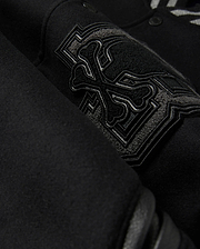 A detailed close-up of the MENS BRAYDEN WOOL LEATHER VARSITY BOMBER - B6J876D200 by Psycho Bunny showcases a black wool-blend fabric with an intricate embroidered design featuring crossbones and decorative elements. The raised and detailed stitching adds texture against the smooth background, reflecting the exceptional craftsmanship typical of a varsity bomber.