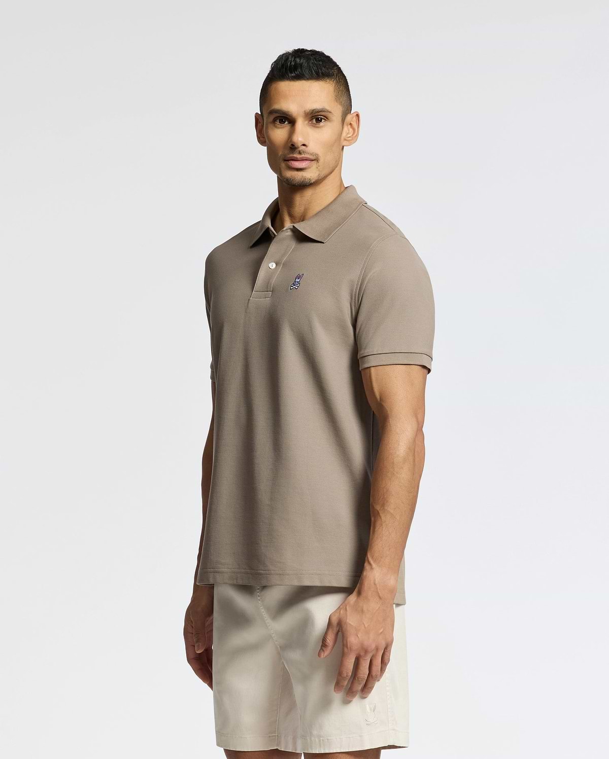 A man stands against a plain background, wearing a Psycho Bunny MENS CLASSIC PIQUE POLO SHIRT - B6K001CRPC with a small chest logo and light beige shorts. He faces forward with a neutral expression.