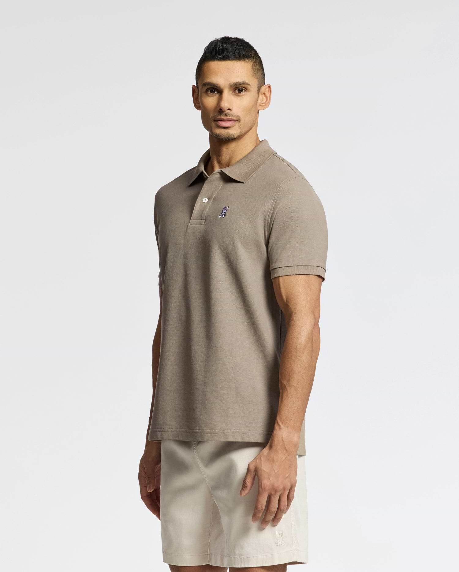 A man stands against a plain background, wearing a Psycho Bunny MENS CLASSIC PIQUE POLO SHIRT - B6K001CRPC with a small chest logo and light beige shorts. He faces forward with a neutral expression.