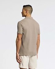 A man faces away, wearing the Psycho Bunny MENS CLASSIC PIQUE POLO SHIRT - B6K001CRPC in taupe Pima cotton, paired with light beige shorts, set against a plain light background.