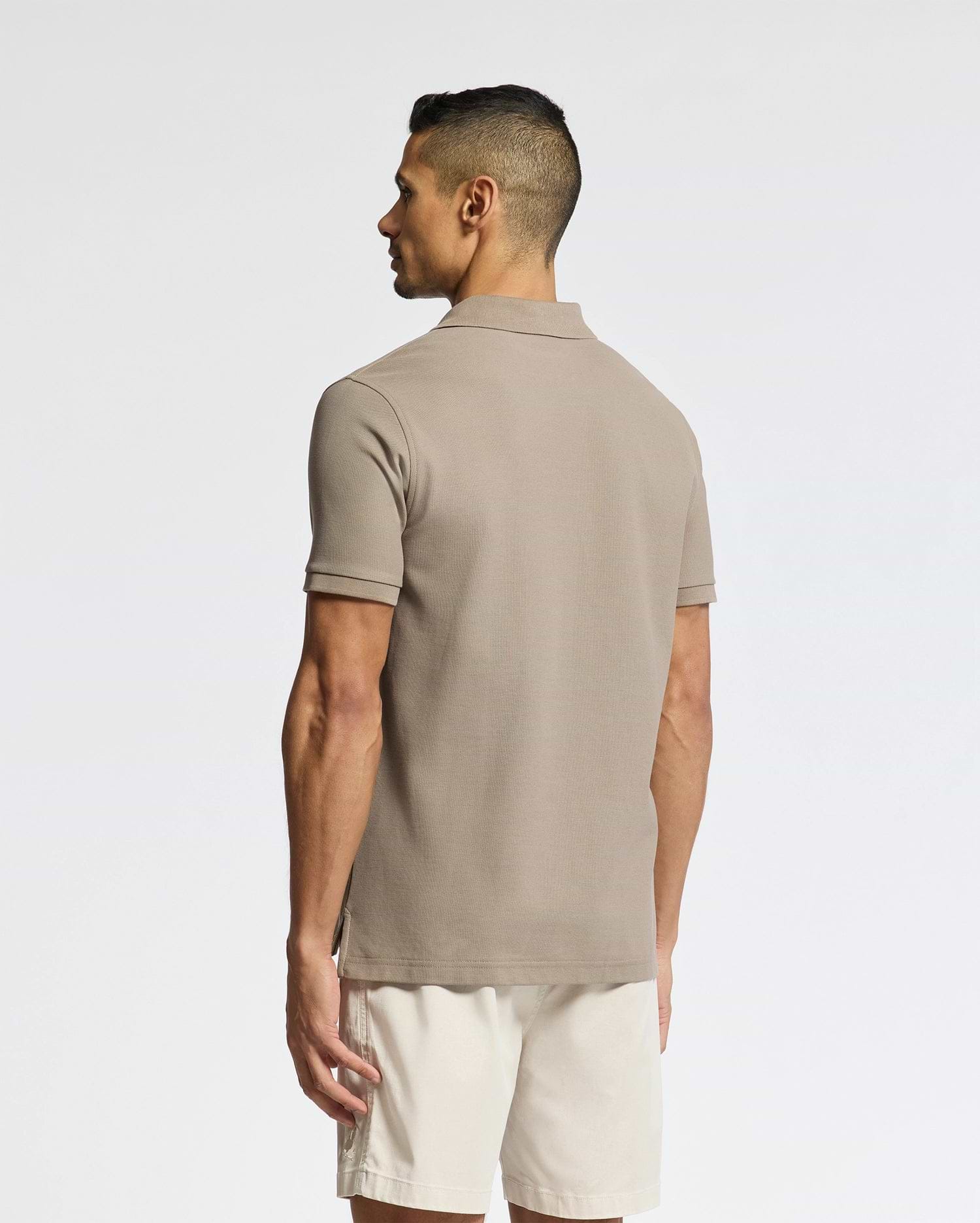 A man faces away, wearing the Psycho Bunny MENS CLASSIC PIQUE POLO SHIRT - B6K001CRPC in taupe Pima cotton, paired with light beige shorts, set against a plain light background.
