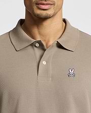 A man wears a taupe Psycho Bunny MENS CLASSIC PIQUE POLO SHIRT (B6K001CRPC) with a collar and buttons, made from soft Pima cotton. A pink bunny and white skull-and-crossbones patch is on the left chest. His face is partially visible.