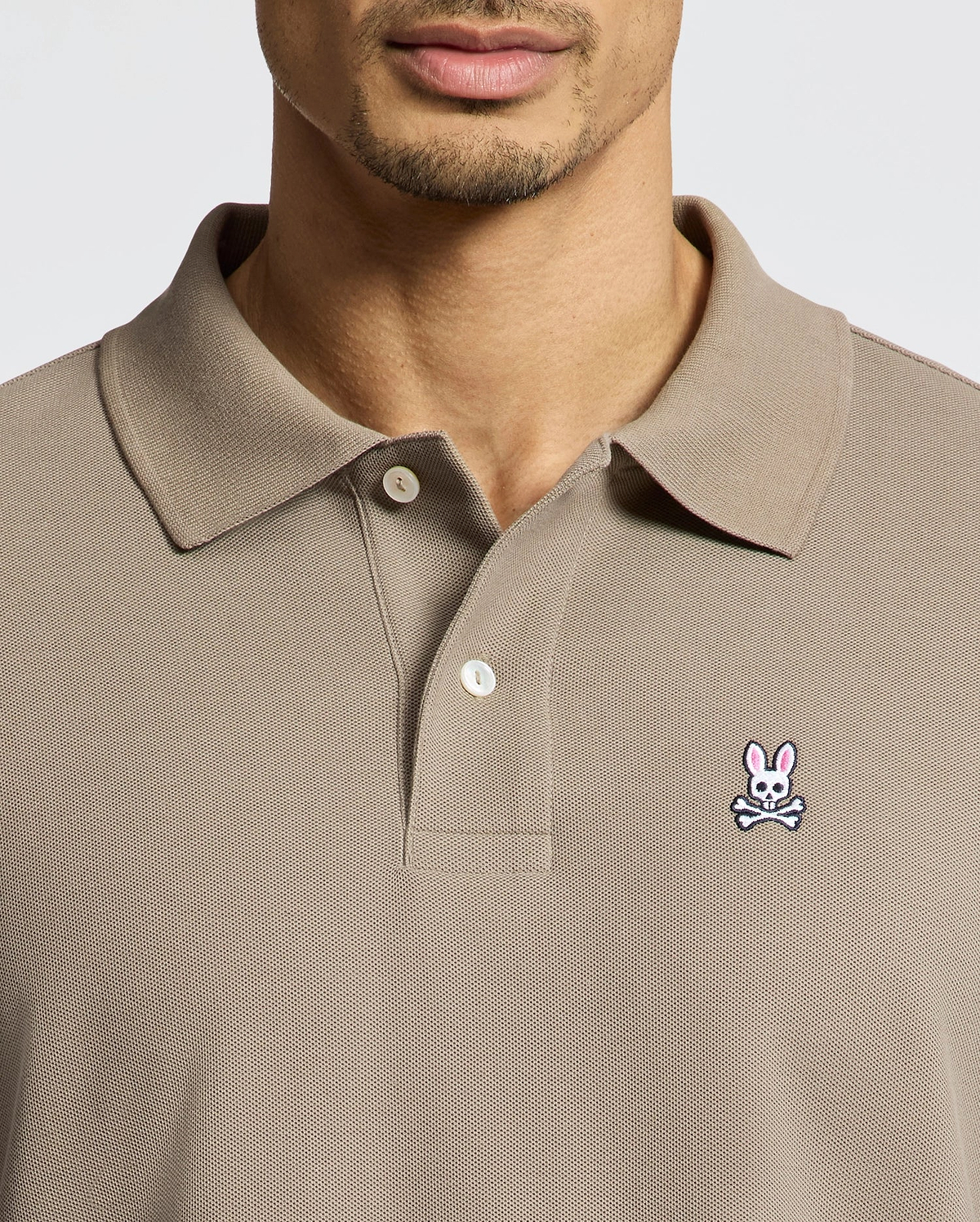 A man wears a taupe Psycho Bunny MENS CLASSIC PIQUE POLO SHIRT (B6K001CRPC) with a collar and buttons, made from soft Pima cotton. A pink bunny and white skull-and-crossbones patch is on the left chest. His face is partially visible.