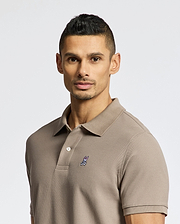 A man with short dark hair and light facial hair wears a Psycho Bunny MENS CLASSIC PIQUE POLO SHIRT - B6K001CRPC in taupe, featuring a small embroidered bunny head on the chest, standing against a plain light background.