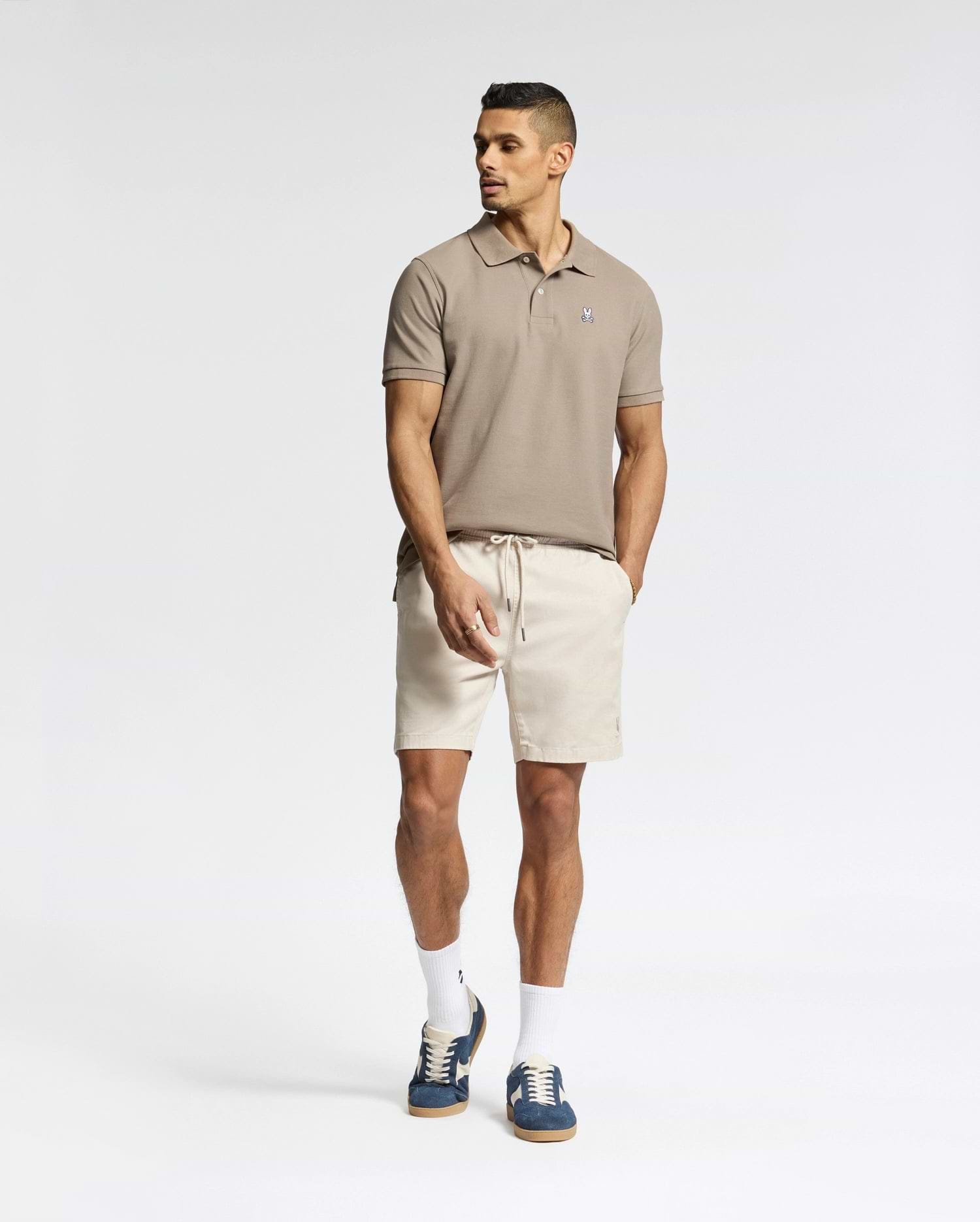 A man stands against a white background wearing a Psycho Bunny MENS CLASSIC PIQUE POLO SHIRT (B6K001CRPC) in beige, paired with light beige shorts, white socks, and navy sneakers. His hands are in his pockets as he looks to the side.