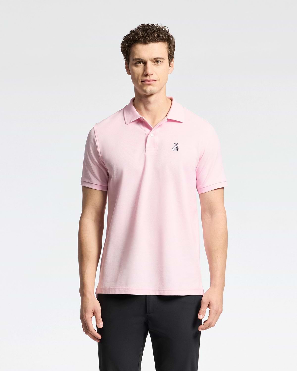 A man stands against a white background, wearing Psycho Bunny's light pink Mens Classic Pique Polo Shirt (B6K001CRPC) made of Peruvian pima cotton with a small chest emblem. He has curly hair, looks directly at the camera, and is also dressed in black pants.