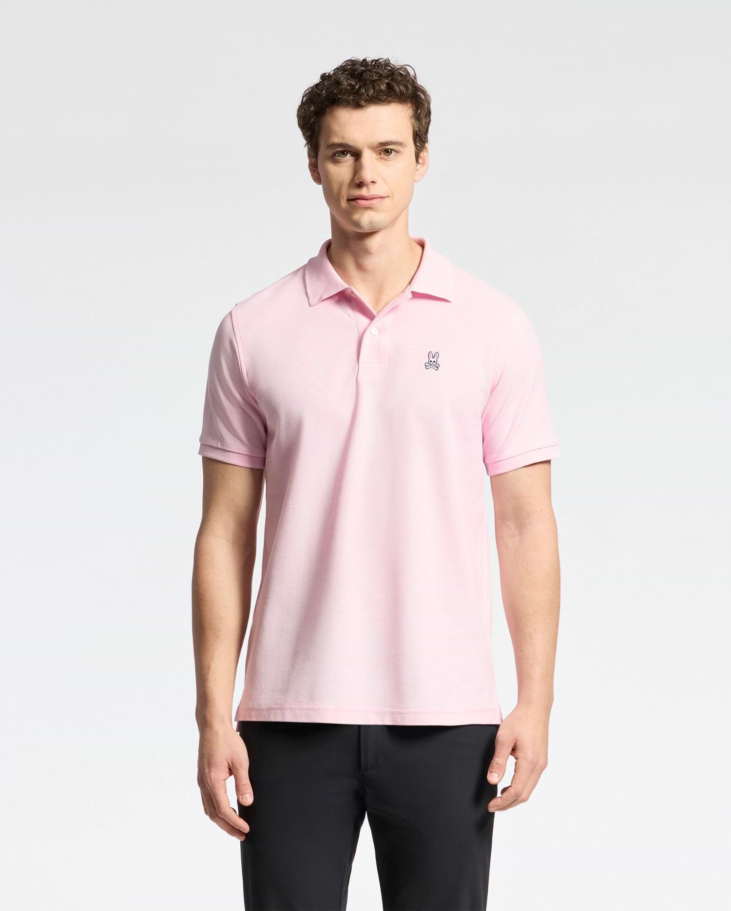 A man stands against a white background, wearing Psycho Bunny's light pink Mens Classic Pique Polo Shirt (B6K001CRPC) made of Peruvian pima cotton with a small chest emblem. He has curly hair, looks directly at the camera, and is also dressed in black pants.