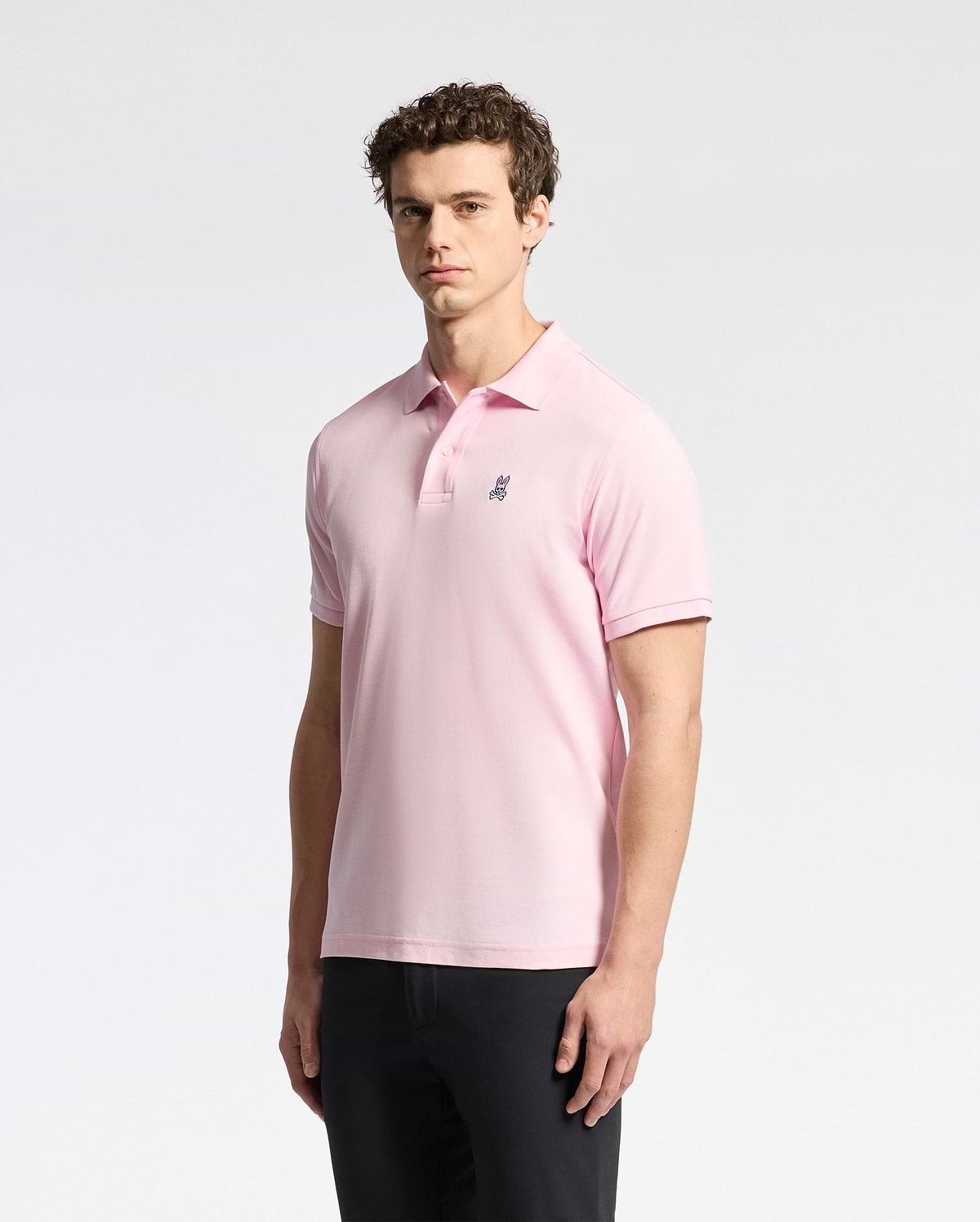 A man wears a Psycho Bunny MENS CLASSIC PIQUE POLO SHIRT (B6K001CRPC) in light pink Peruvian pima cotton, with an embroidered logo on the chest, paired with black pants against a plain white background.