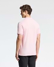 A curly-haired man is seen from behind, donning the Psycho Bunny light pink MENS CLASSIC PIQUE POLO SHIRT - B6K001CRPC in luxurious pima cotton with black pants, against a plain light backdrop.