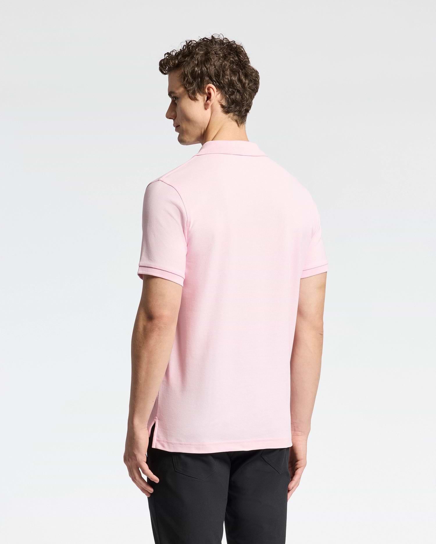 A curly-haired man is seen from behind, donning the Psycho Bunny light pink MENS CLASSIC PIQUE POLO SHIRT - B6K001CRPC in luxurious pima cotton with black pants, against a plain light backdrop.