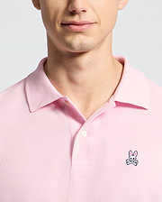 A person is wearing the Psycho Bunny MENS CLASSIC PIQUE POLO SHIRT - B6K001CRPC in pink, featuring a bunny and crossbones logo on the left chest. The photo highlights their upper chest and partially visible face against a simple light background.
