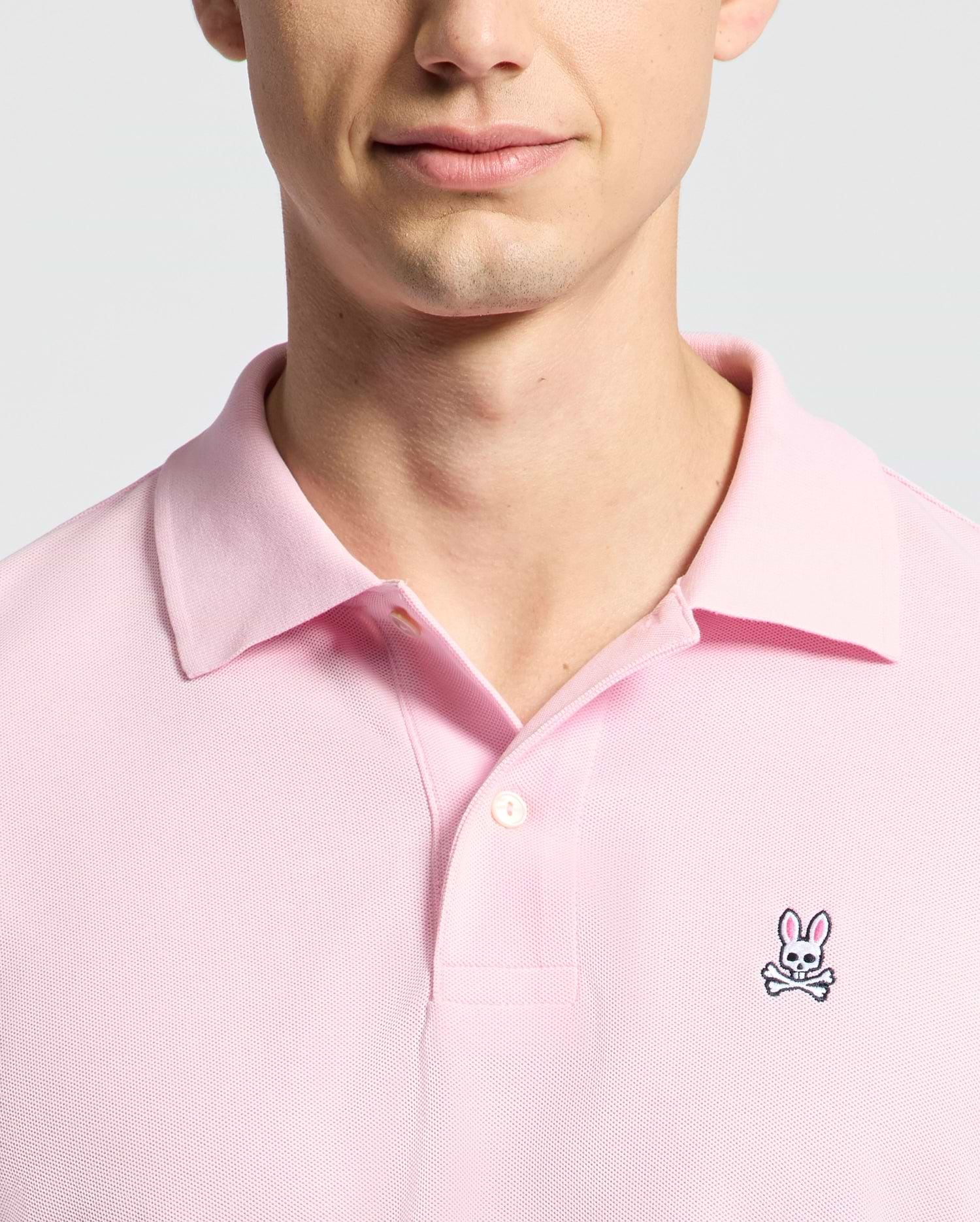 A person is wearing the Psycho Bunny MENS CLASSIC PIQUE POLO SHIRT - B6K001CRPC in pink, featuring a bunny and crossbones logo on the left chest. The photo highlights their upper chest and partially visible face against a simple light background.