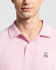 A person is wearing the Psycho Bunny MENS CLASSIC PIQUE POLO SHIRT - B6K001CRPC in pink, featuring a bunny and crossbones logo on the left chest. The photo highlights their upper chest and partially visible face against a simple light background.