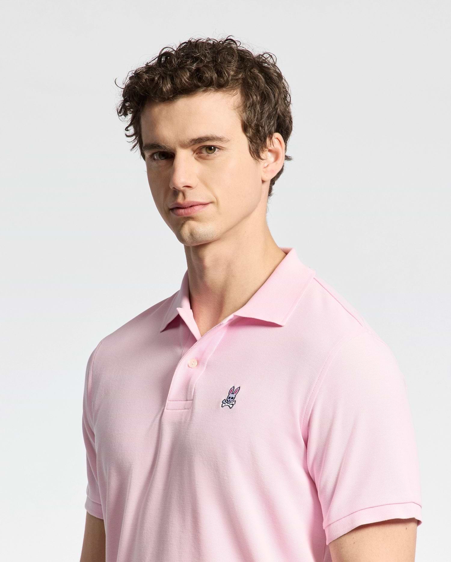 A person with curly hair wears a Psycho Bunny MENS CLASSIC PIQUE POLO SHIRT - B6K001CRPC in light pink, made of luxurious Peruvian pima cotton, featuring a small embroidered bunny logo. They stand against a plain gray background, looking slightly to the side.