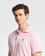 A person with curly hair wears a Psycho Bunny MENS CLASSIC PIQUE POLO SHIRT - B6K001CRPC in light pink, made of luxurious Peruvian pima cotton, featuring a small embroidered bunny logo. They stand against a plain gray background, looking slightly to the side.