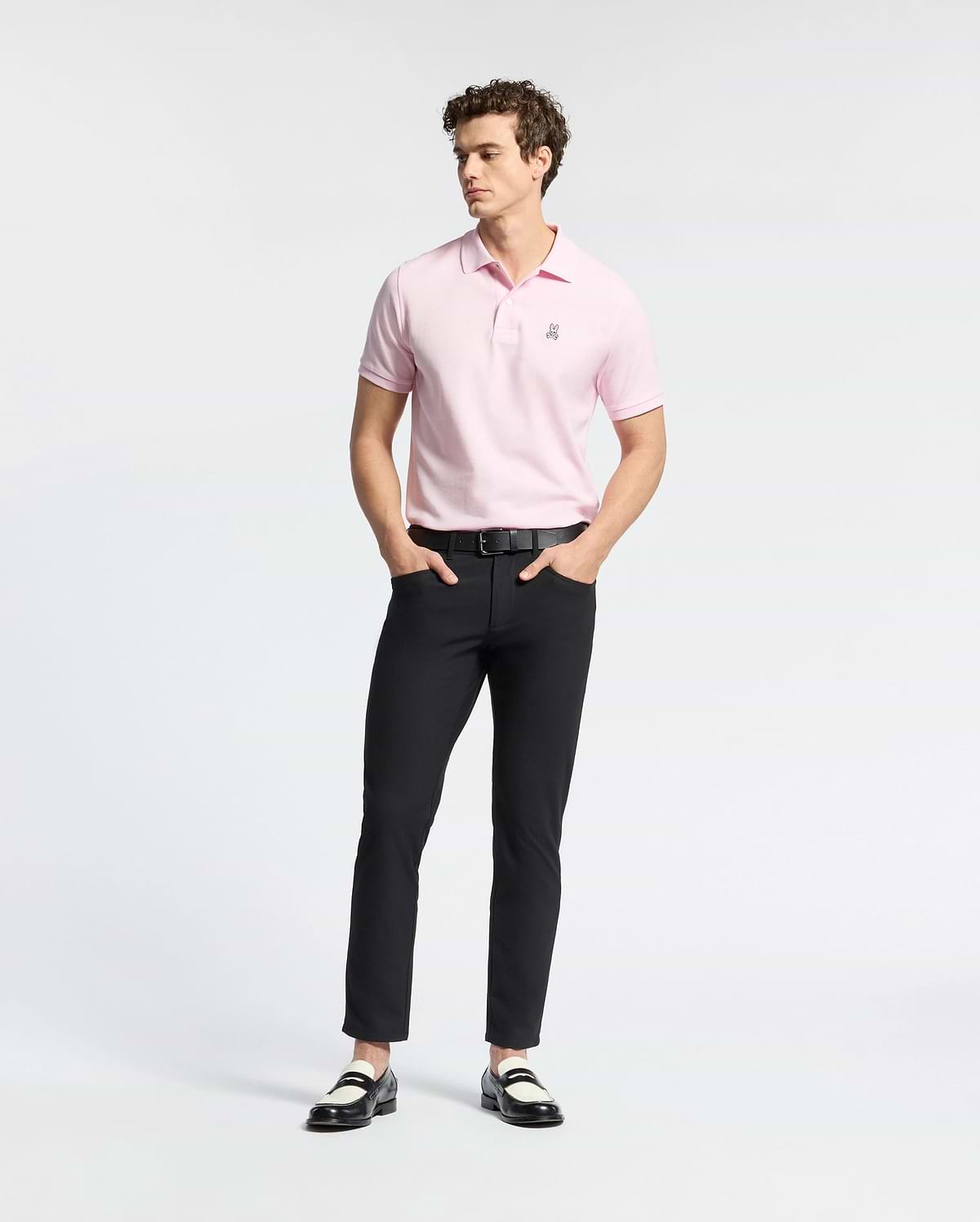 A person wearing a light pink Psycho Bunny MENS CLASSIC PIQUE POLO SHIRT - B6K001CRPC, black pants, and black and white loafers stands against a plain white background. They have curly hair and their hands are in their pockets.