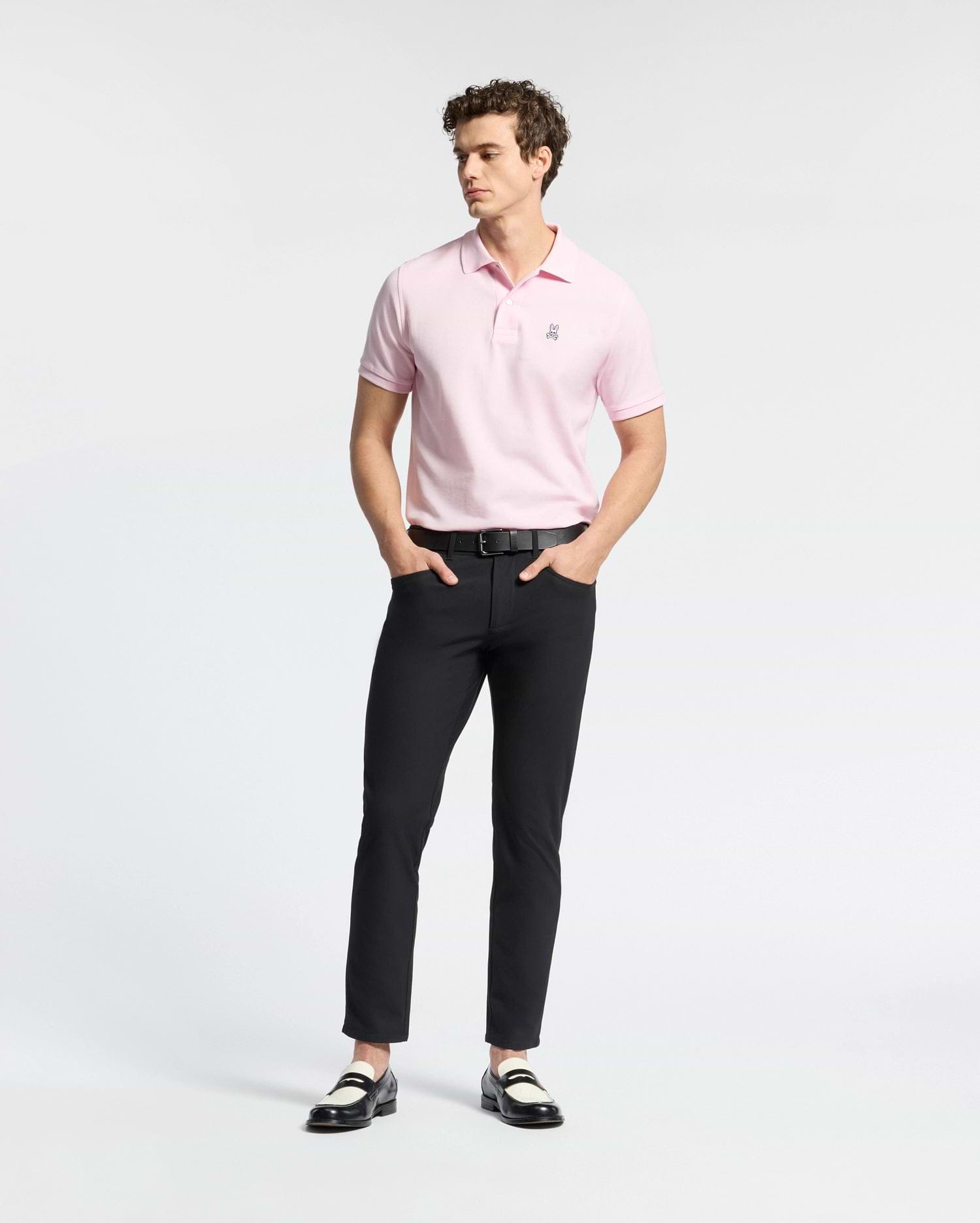 A person wearing a light pink Psycho Bunny MENS CLASSIC PIQUE POLO SHIRT - B6K001CRPC, black pants, and black and white loafers stands against a plain white background. They have curly hair and their hands are in their pockets.