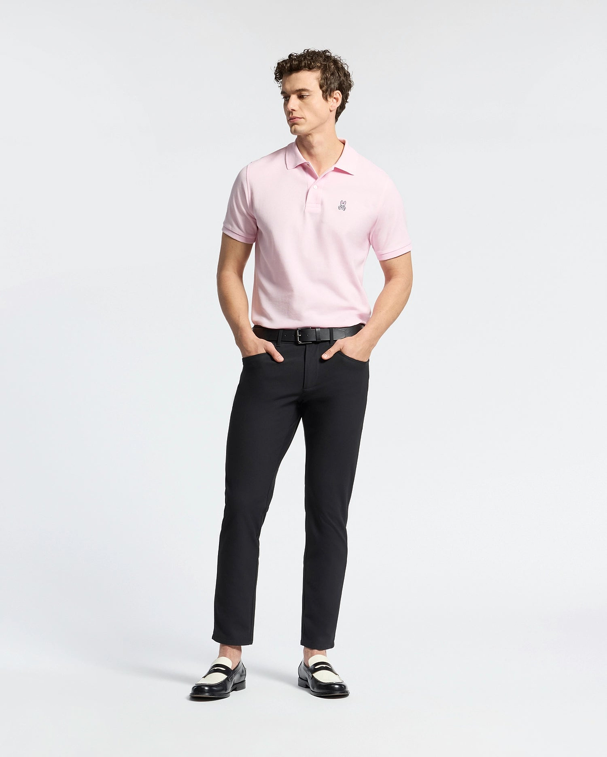 A person wearing a light pink Psycho Bunny MENS CLASSIC PIQUE POLO SHIRT - B6K001CRPC, black pants, and black and white loafers stands against a plain white background. They have curly hair and their hands are in their pockets.