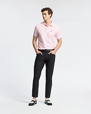 A person wearing a light pink Psycho Bunny MENS CLASSIC PIQUE POLO SHIRT - B6K001CRPC, black pants, and black and white loafers stands against a plain white background. They have curly hair and their hands are in their pockets.
