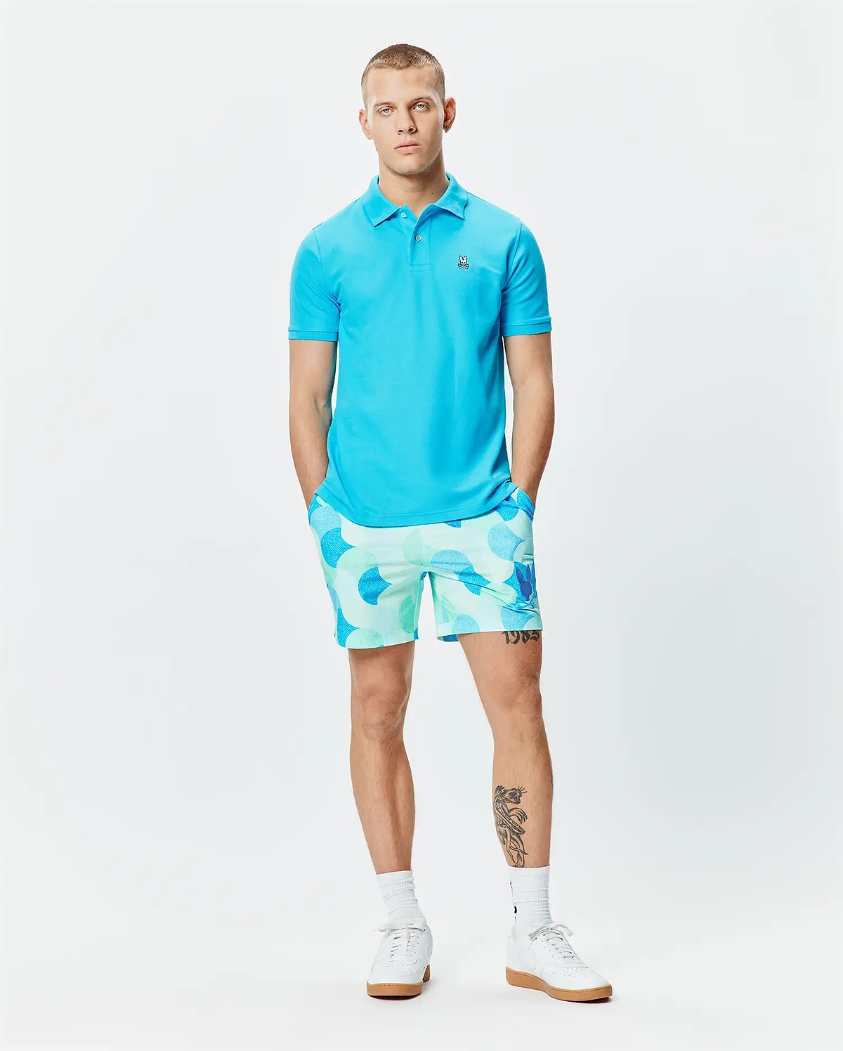 Mens Classic Pique Polo in vibrant cyan, featuring a classic fit and small embroidered Psycho Bunny skull logo on the left chest. Paired with relaxed-fit shorts in a light blue and mint green all-over circular pattern. (6)