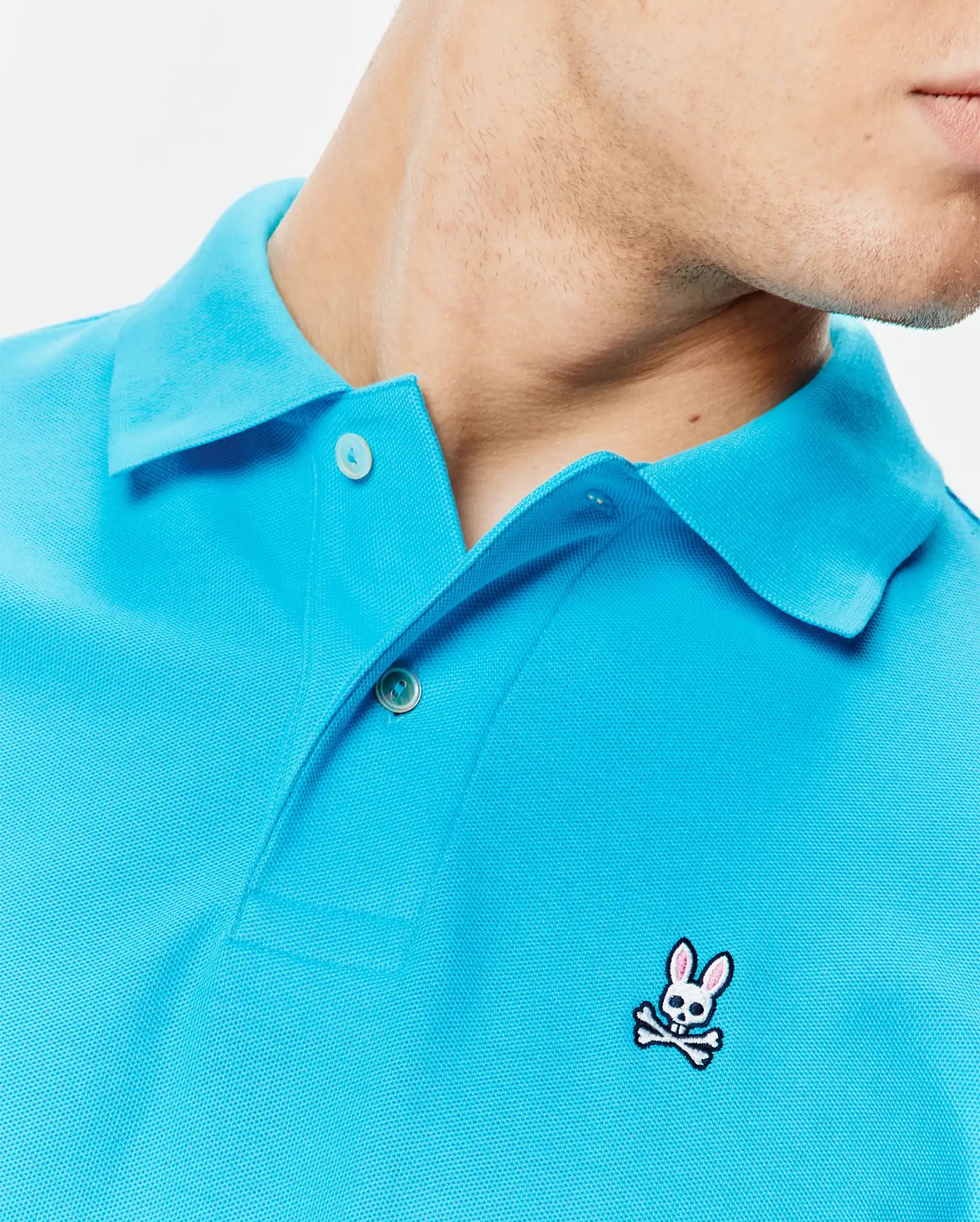 Mens Classic Pique Polo in bright aqua blue, featuring a textured pique knit, two-button placket with white buttons, and a small embroidered white and pink Psycho Bunny skull logo on the left chest. (4)